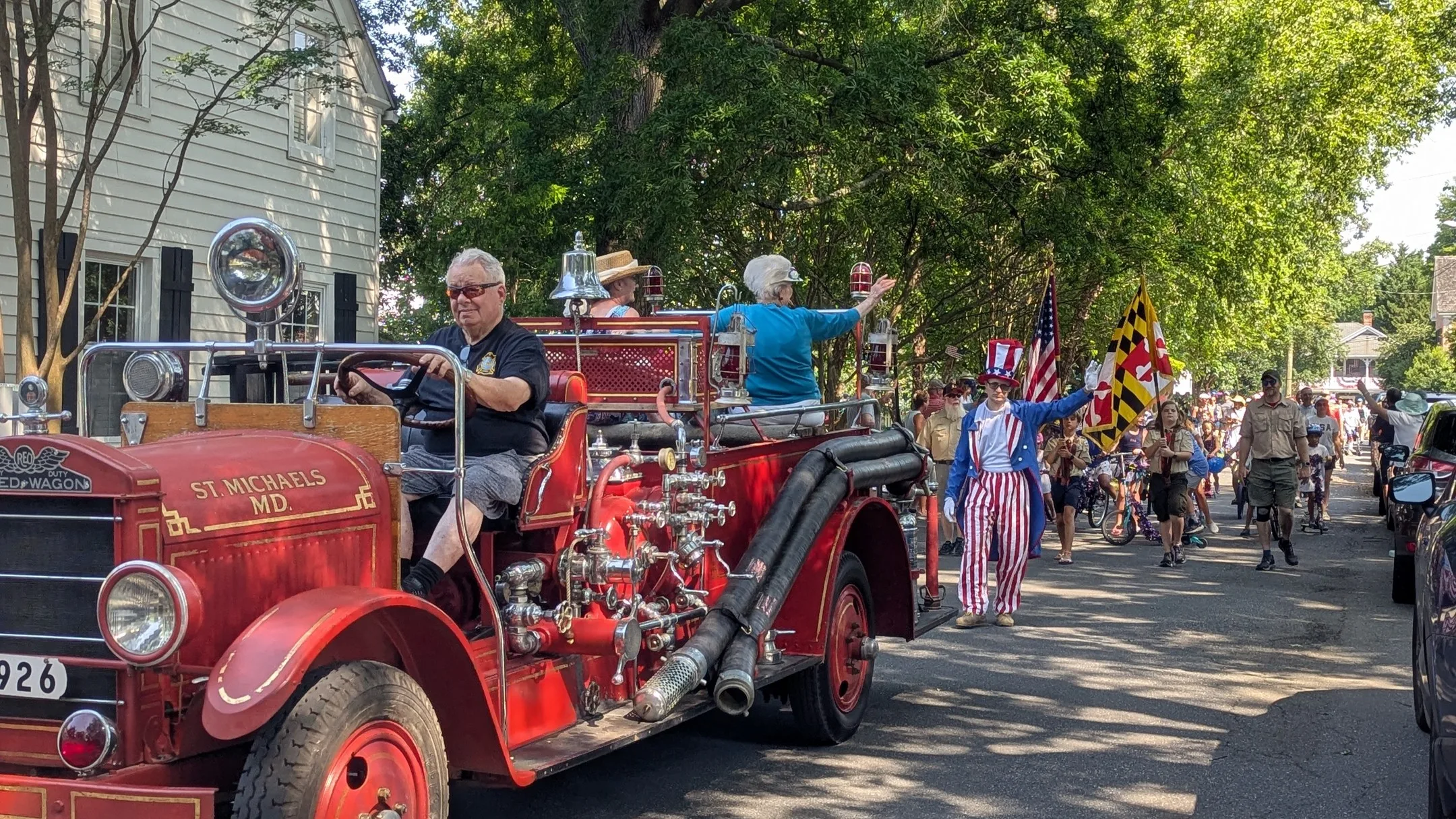 Street parade led by an old-fashioned fire truck with an American and Maryland flag in the background.