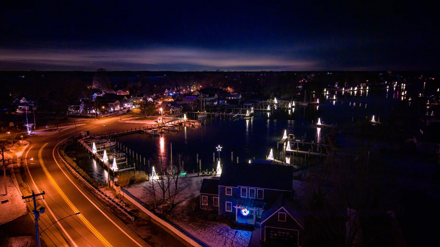 Road leading into town with water to the right, showing lighted Christmas trees on town docks.