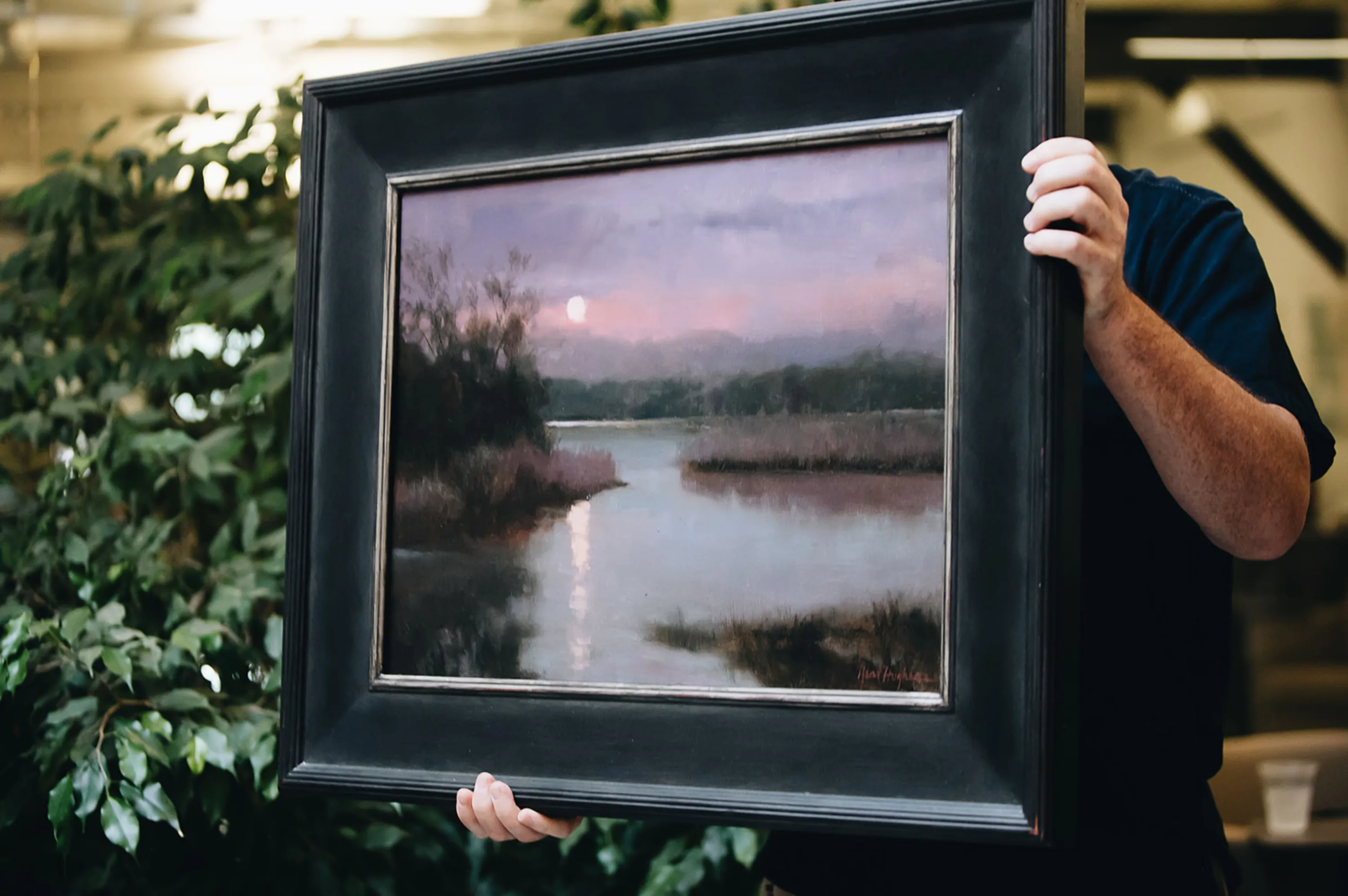 Man holding a framed watercolor painting of a winding tidal river view with a setting sun.