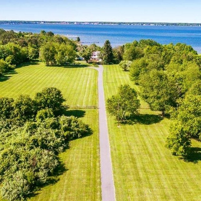 Green field with trees on either side, and a road in the middle leading to a large, waterfront home.