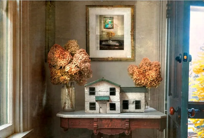 Room with art work on the wall above a table with a wooden model house flanked by flowers on either side.